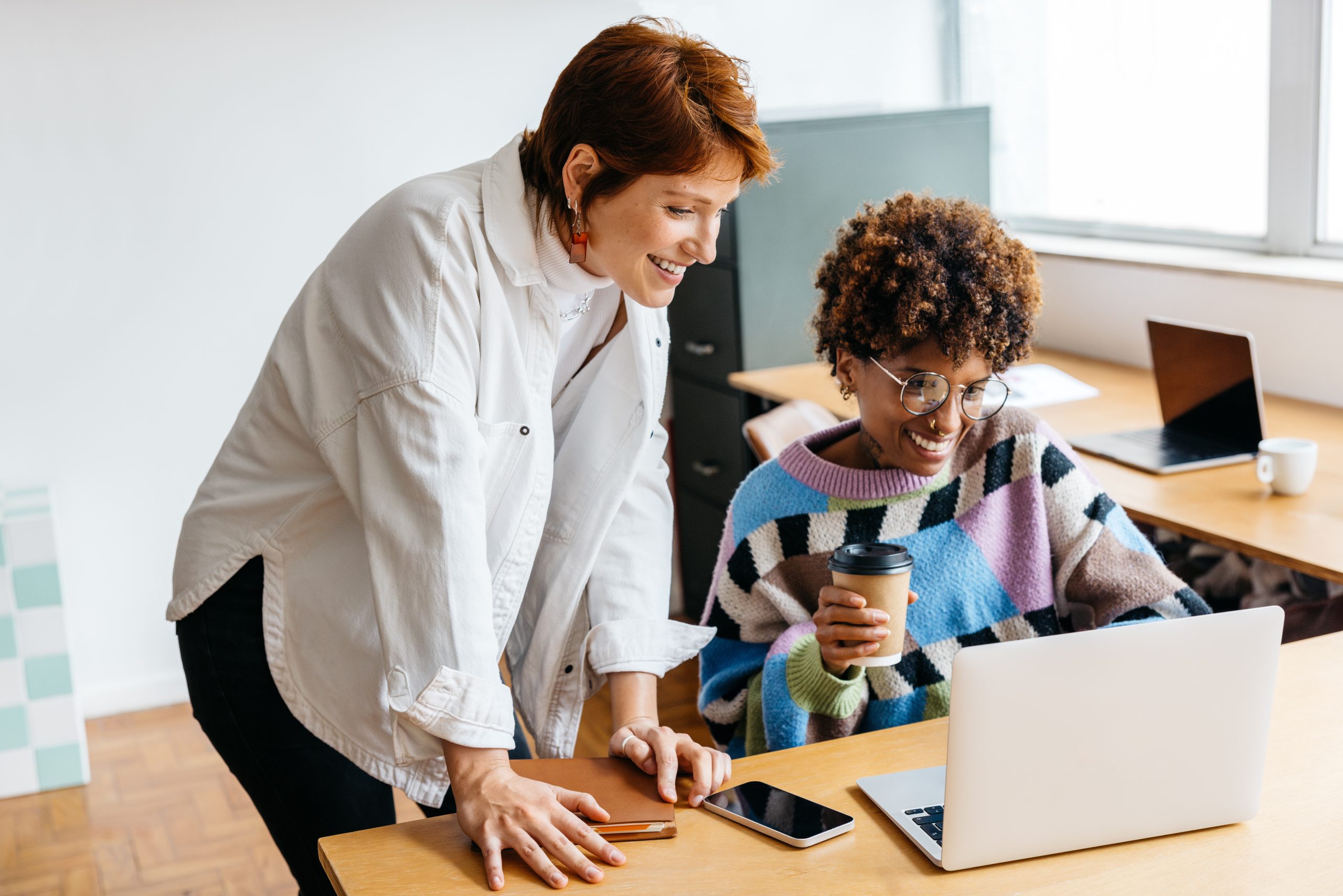 Two diverse colleagues are engaged in a project in a co-working space. One stands with a coffee cup, smiling at a laptop screen, demonstrating a positive, collaborative work environment with technology aiding their freelance work.